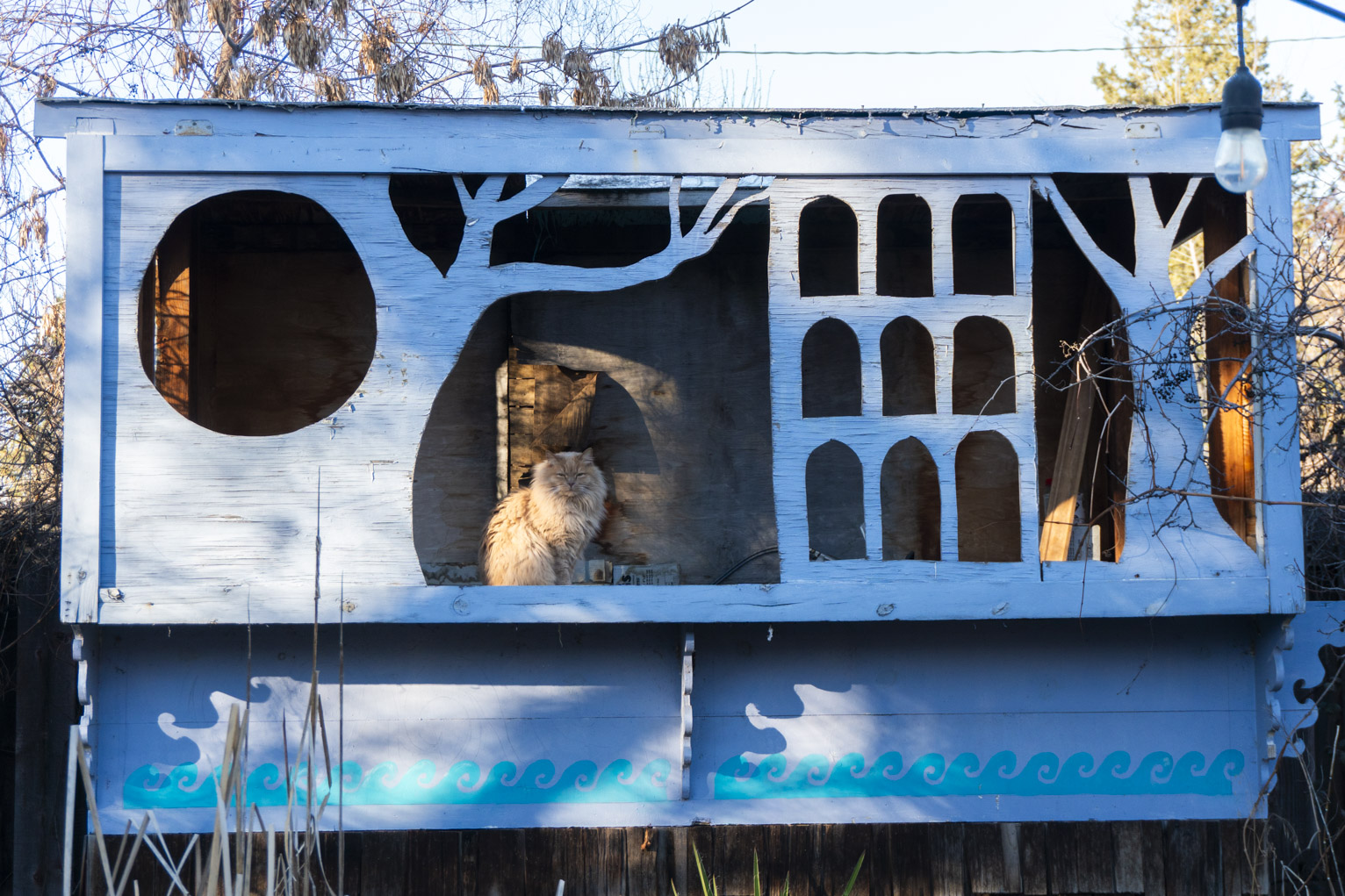 A cat sits in a blue wood structure with cutouts of trees and windows